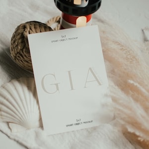 Puede incluir: Una tarjeta blanca con el texto "GIA" en una fuente serif. La tarjeta mide 12,7 x 17,8 cm y es una maqueta de objeto inteligente. La tarjeta está sobre una superficie blanca con una concha, una bola de cuerda y una pequeña figura de faro.
