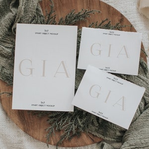 Peut inclure: Trois cartes blanches avec le mot "GIA" imprim&eacute; en brun clair. Les cartes sont de tailles diff&eacute;rentes et portent l'inscription "SMART OBJECT MOCKUP" ainsi que les dimensions. Les cartes sont pos&eacute;es sur une surface en bois avec de la verdure et un tissu vert.