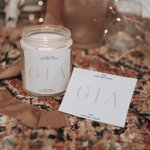 Op de afbeelding: Een witte kaars pot met een label waarop "GIA" en "CANDLE LABEL SMART OBJECT MOCKUP" staat. De kaars pot staat op een bruin en oranje gestreept tapijt. Er ligt een witte kaart met hetzelfde label en tekst op het tapijt naast de kaars pot.