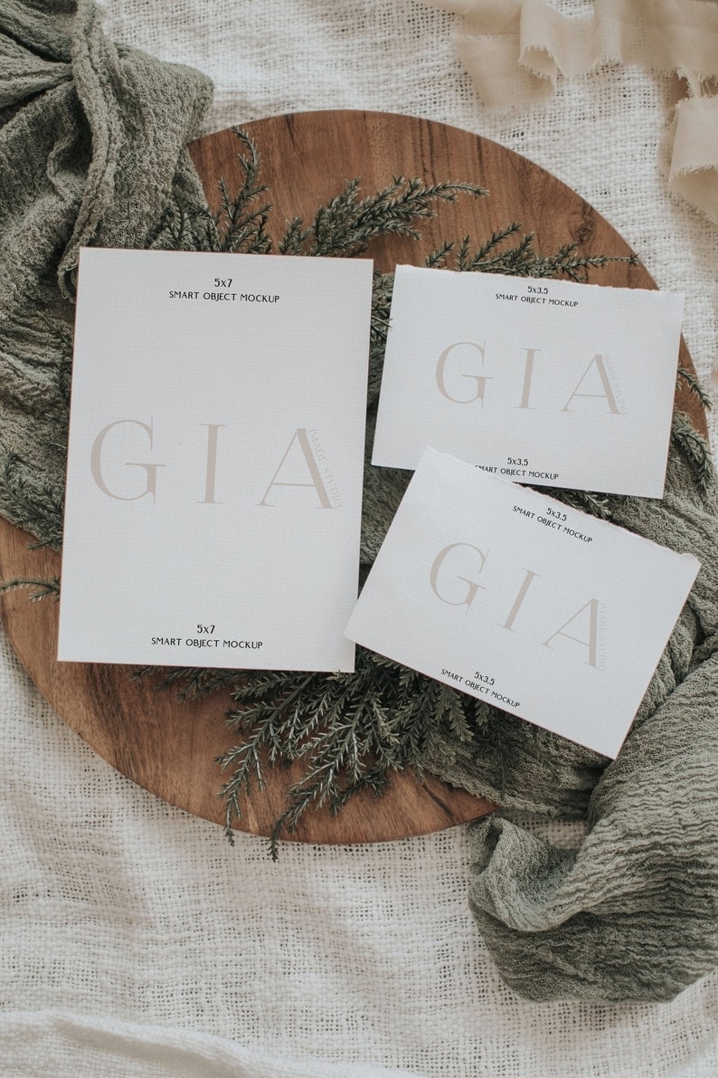 Peut inclure: Trois cartes blanches avec le mot "GIA" imprim&eacute; en brun clair. Les cartes sont de tailles diff&eacute;rentes et portent l'inscription "SMART OBJECT MOCKUP" ainsi que les dimensions. Les cartes sont pos&eacute;es sur une surface en bois avec de la verdure et un tissu vert.