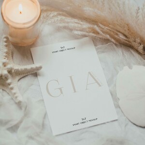 May include: A white card with the text "GIA" in a serif font and the text "5x7 SMART OBJECT MOCKUP" at the top and bottom of the card. The card is lying on a white surface with a white candle, a starfish, and a dried pampas grass.