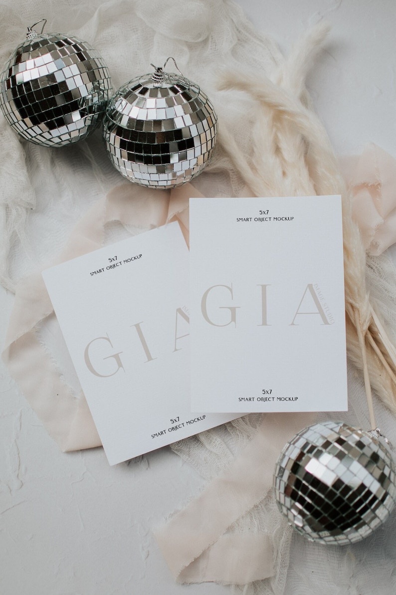 May include: Two white cards with the text "5x7 SMART OBJECT MOCKUP" and the word "GIAGIA" in a light brown font. The cards are on a white surface with a white feather plume and three silver disco balls.
