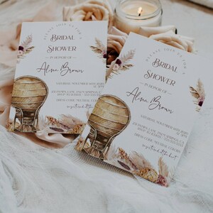 May include: Two white cards with a boho floral design and a wicker chair illustration. The text on the cards reads "Please join us for a bridal shower in honor of Alina Brown" and includes the date, time, and location of the event.