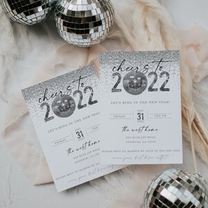 May include: A silver glitter New Year's Eve party invitation with the text "cheer's to 2022" and a disco ball graphic. The invitation includes the date, time, and location of the party. The text "Let's ring in the new year!" is also included.