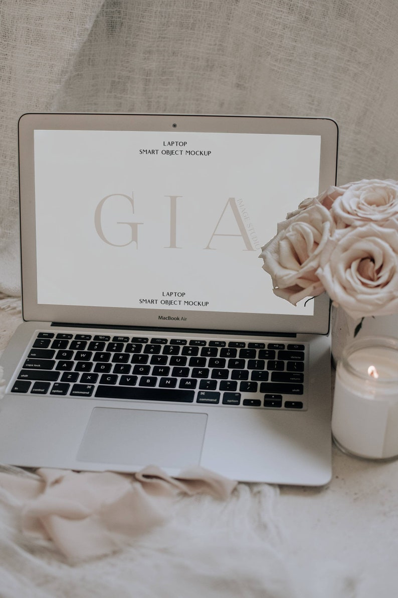 May include: A silver laptop computer with a white screen displaying the text "GIA" and the words "Laptop Smart Object Mockup". The laptop is sitting on a white surface with a bouquet of light pink roses and a white candle in a glass jar.