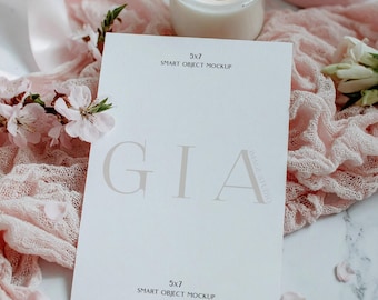 May include: A 5x7 inch white card mockup with the name "GIA" printed in a light brown color. The card is surrounded by pink flowers, a lit candle in a glass jar, and pink ribbons on a marble surface.
