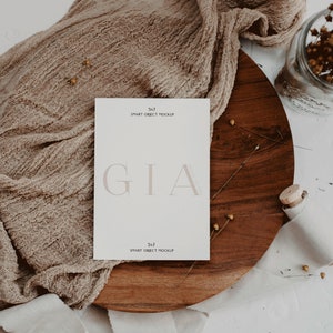 May include: A white card with the text "GIA" in a light brown font. The card is on a wooden surface with a brown fabric draped over it. The text "5x7 SMART OBJECT MOCKUP" appears at the top and bottom of the card.