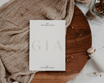 May include: A white card with the text "GIA" in a light brown font. The card is on a wooden surface with a brown fabric draped over it. The text "5x7 SMART OBJECT MOCKUP" appears at the top and bottom of the card.