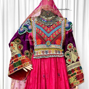 Könnte beinhalten: Ein traditionelles rosa Kleid mit bunten Stickereien und Perlen. Das Kleid hat lange Ärmel mit aufwendigen Details und ein passendes Kopftuch. Das Kleid ist mit einer silbernen Halskette mit bunten Steinen verziert.