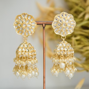 May include: A pair of gold earrings with a floral design and pearl accents. The earrings are hanging from a gold stand.