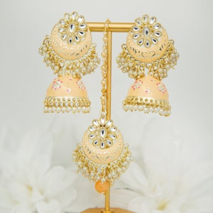 May include: A pair of peach-colored jhumka earrings with gold accents and pearl detailing. The earrings are displayed on a gold stand.