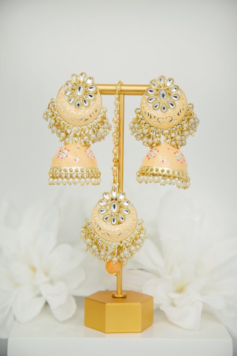 May include: A pair of peach-colored jhumka earrings with gold accents and pearl detailing. The earrings are displayed on a gold stand.