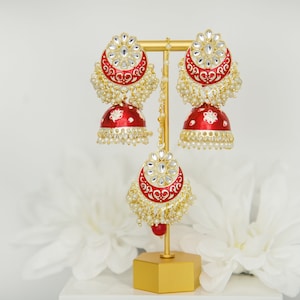 May include: A pair of red and gold Indian earrings with white pearl accents. The earrings are on a gold stand.