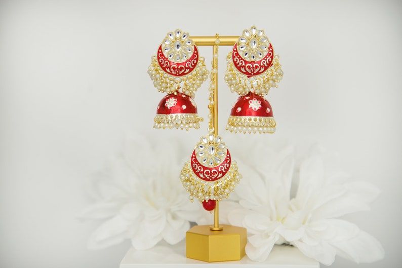 May include: A pair of red and gold Indian earrings with white pearl accents. The earrings are on a gold stand.