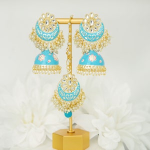 May include: A pair of turquoise blue jhumka earrings with gold accents and pearl detailing. The earrings are displayed on a gold stand.