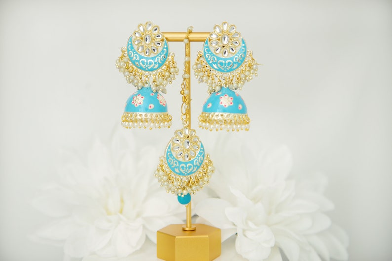 May include: A pair of turquoise blue jhumka earrings with gold accents and pearl detailing. The earrings are displayed on a gold stand.