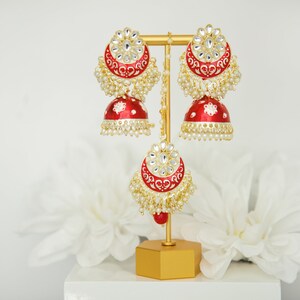 May include: A pair of red and gold earrings with white stones and pearl accents. The earrings are on a gold stand.