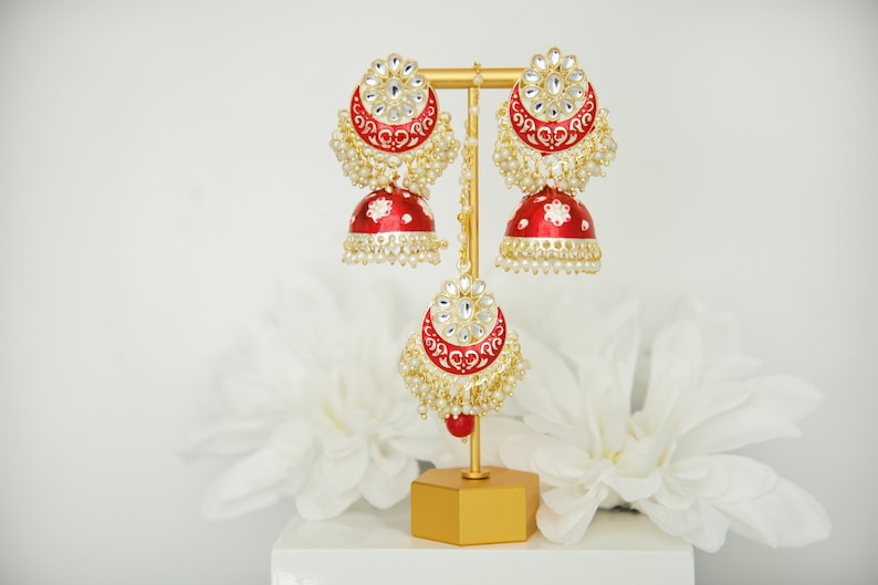 May include: A pair of red and gold earrings with white stones and pearl accents. The earrings are on a gold stand.