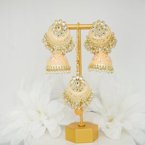 May include: A pair of peach-colored earrings with gold accents and pearl embellishments. The earrings are on a gold stand with a peach-colored tassel.