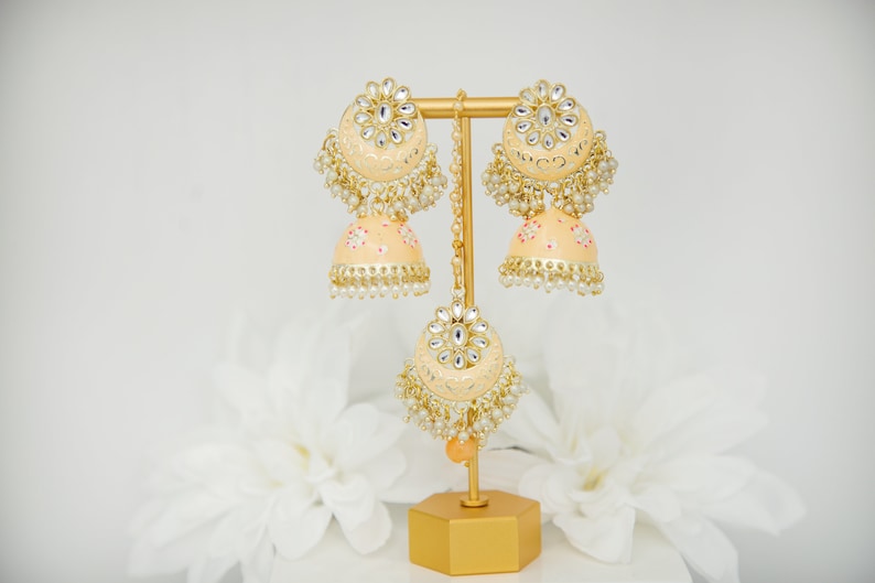 May include: A pair of peach-colored earrings with gold accents and pearl embellishments. The earrings are on a gold stand with a peach-colored tassel.