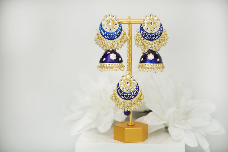 May include: A pair of blue and gold earrings with pearl accents. The earrings are on a gold stand. The earrings are a traditional Indian style.