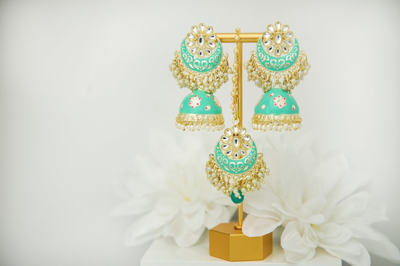 May include: A pair of teal blue Indian earrings with gold accents and pearl embellishments. The earrings are displayed on a gold stand.