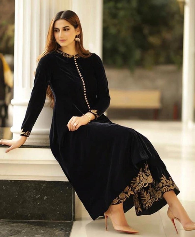 May include: A long, black velvet dress with gold embroidery on the sleeves and hem. The dress features a row of gold buttons down the front and is paired with nude heels. The outfit is elegant and suitable for formal occasions.