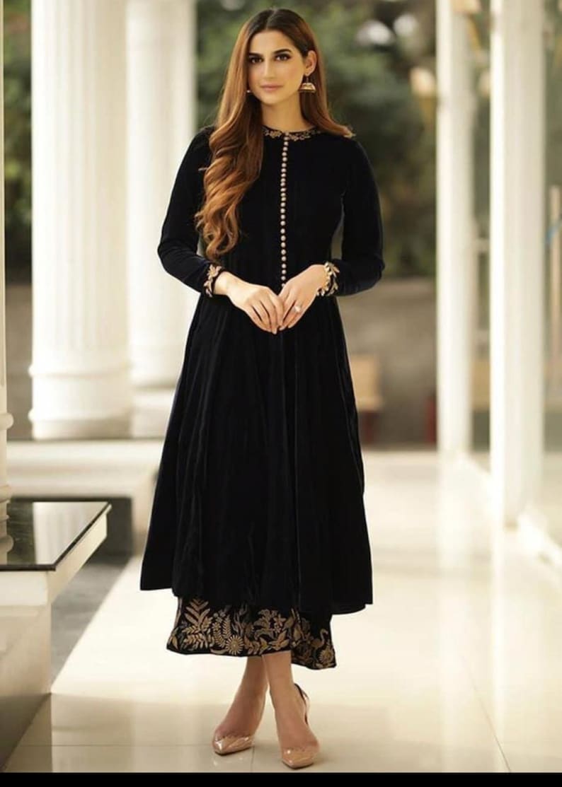 May include: A long, black velvet dress with long sleeves and a button-down front. The dress features gold embroidery on the cuffs and hem. The model is wearing gold earrings and nude heels.