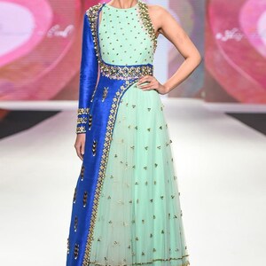 May include: A mint green and blue formal gown with gold embellishments. The dress features a one-shoulder design with a long blue sleeve and intricate gold detailing. The skirt is a flowing, layered design with gold accents.