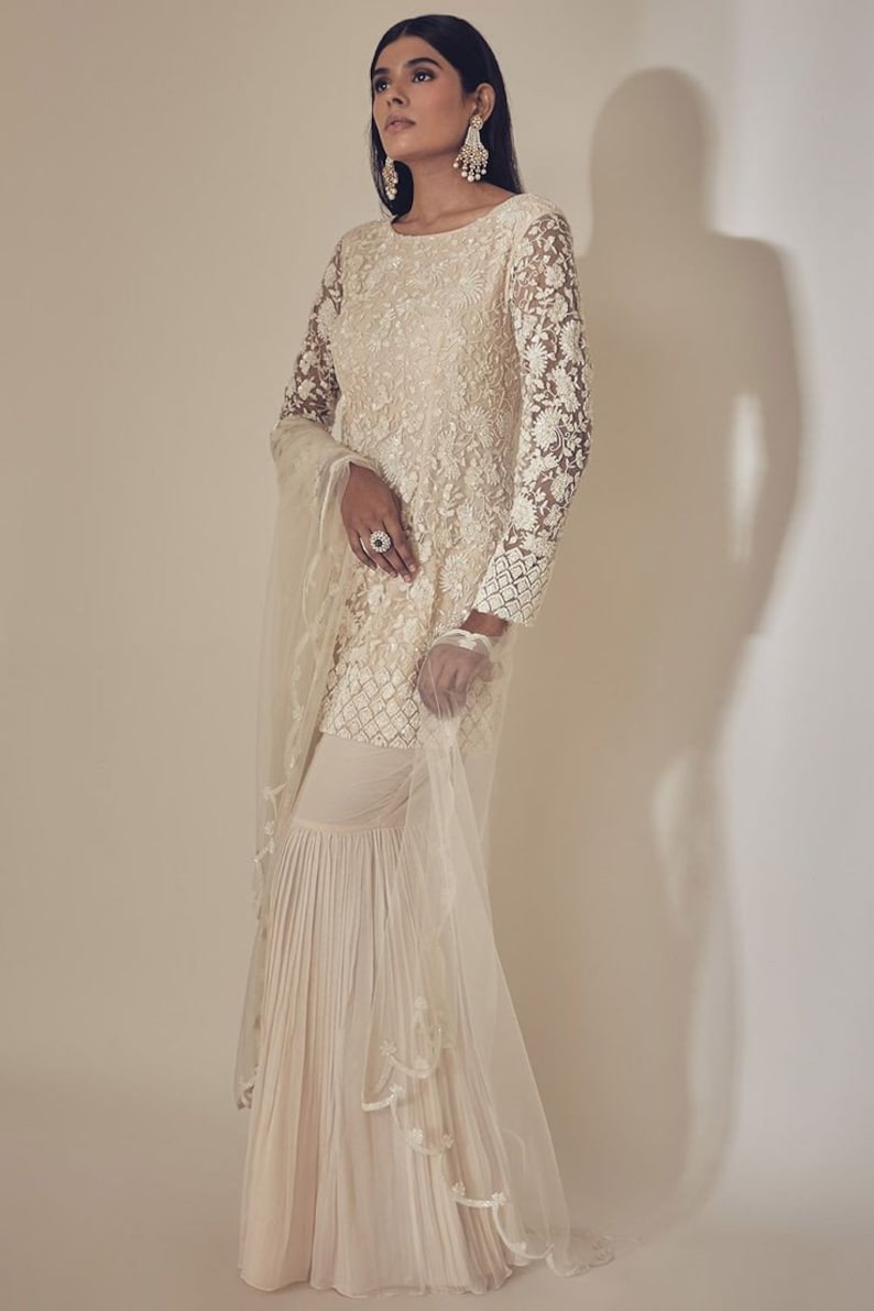 May include: Ivory embroidered Sharara suit. The long-sleeved top has intricate floral lace patterns, paired with matching flared trousers. A sheer dupatta with delicate trim completes the ensemble. Suitable for special occasions.