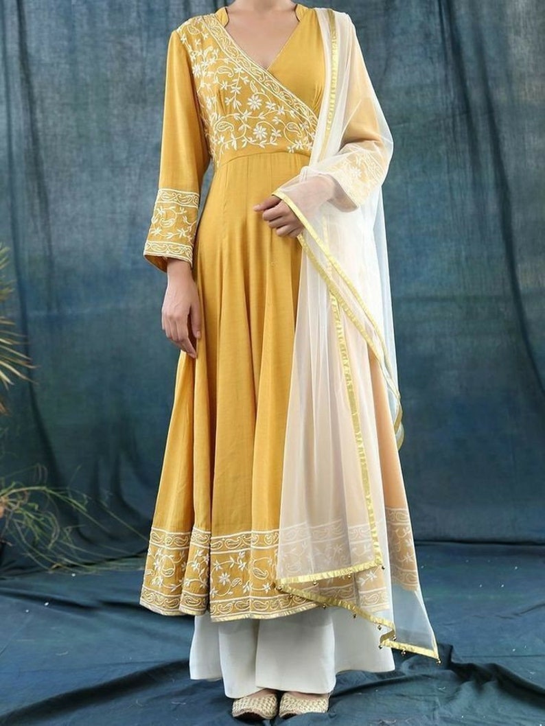 May include: Mustard yellow wrap dress with white floral embroidery on the bodice and sleeves. The dress has a flowing skirt and is paired with white palazzo pants and a sheer white scarf with gold trim. The outfit is elegant and suitable for special occasions.