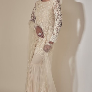 May include: Ivory embroidered Sharara suit. The long-sleeved top has intricate floral lace patterns, paired with matching flared trousers. A sheer dupatta with delicate trim completes the ensemble. Suitable for special occasions.