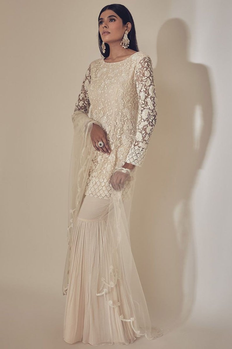 May include: Ivory embroidered Sharara suit. The long-sleeved top has intricate floral lace patterns, paired with matching flared trousers. A sheer dupatta with delicate trim completes the ensemble. Suitable for special occasions.