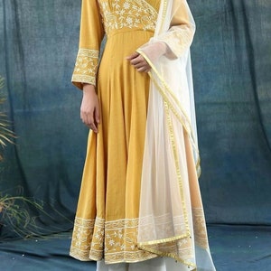 May include: Mustard yellow wrap dress with white floral embroidery on the bodice and sleeves. The dress has long sleeves and a flowing skirt. A sheer white scarf with gold trim is draped over the shoulder. The outfit is paired with white pants.