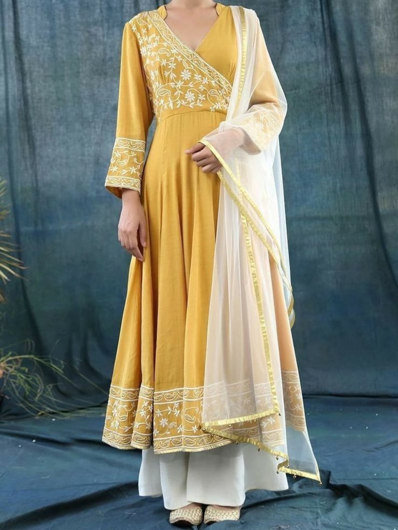 May include: Mustard yellow wrap dress with white floral embroidery on the bodice and sleeves. The dress has long sleeves and a flowing skirt. A sheer white scarf with gold trim is draped over the shoulder. The outfit is paired with white pants.