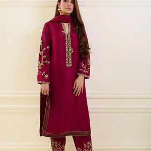 May include: A burgundy-colored, long-sleeved tunic and matching pants set. The tunic features gold embroidery along the neckline, sleeves, and hem. The pants have matching gold embroidery at the hem. A burgundy scarf completes the ensemble.
