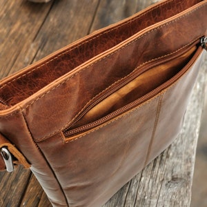 May include: A brown leather crossbody bag with a zippered pocket and a flap closure. The bag is open, revealing the brown fabric lining.