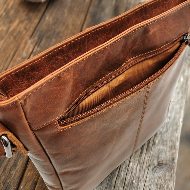 May include: A brown leather crossbody bag with a zippered pocket and a flap closure. The bag is open, revealing the brown fabric lining.