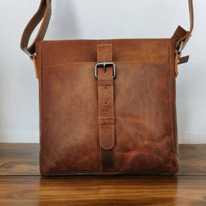 May include: A brown leather satchel bag with a buckle closure and a long adjustable strap. The bag has a distressed look and a worn texture.