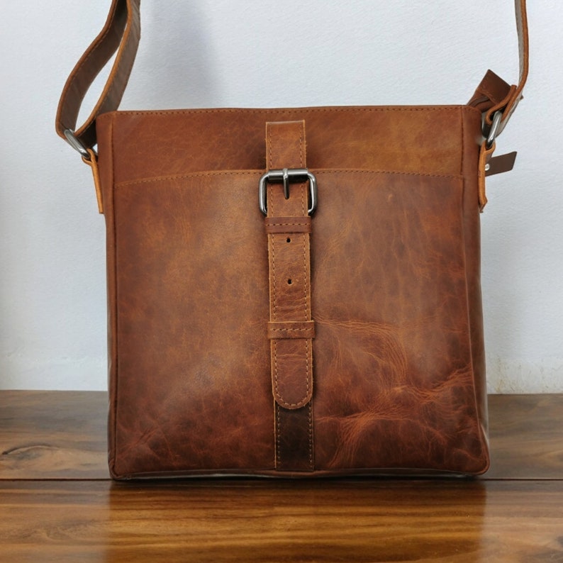 May include: A brown leather satchel bag with a buckle closure and a long adjustable strap. The bag has a distressed look and a worn texture.