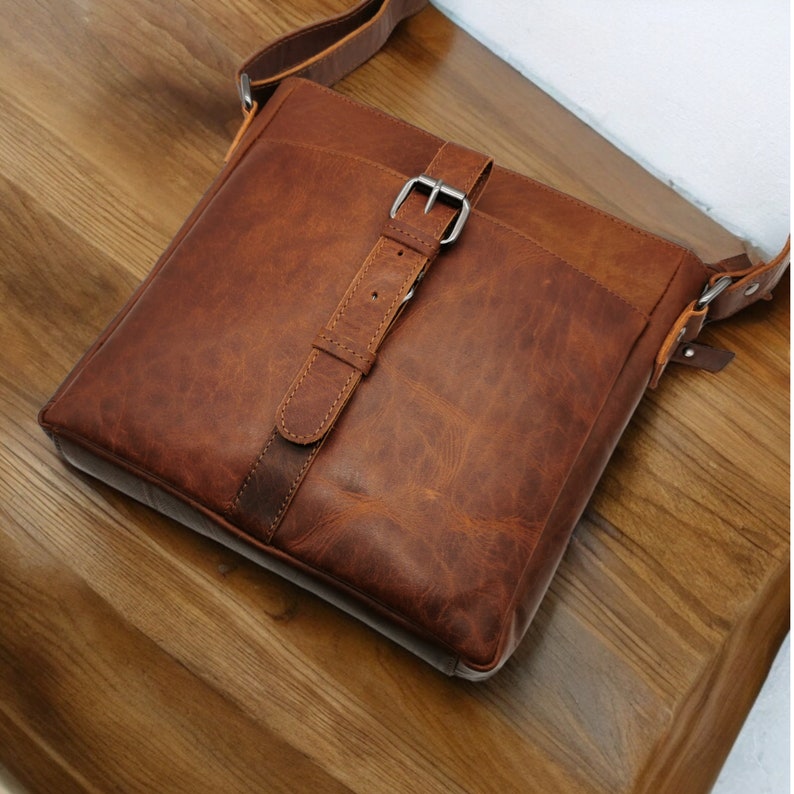 May include: A brown leather messenger bag with a silver buckle and a strap. The bag has a worn, distressed look.