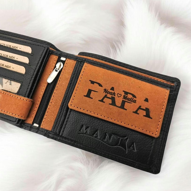 Könnte beinhalten: Eine schwarze Ledergeldbörse mit einer braunen Lederklappe. Die Klappe trägt den gravierten Schriftzug "PAPA", darüber die Namen "Noah" und "Malia" sowie ein Herzsymbol. Die Geldbörse trägt außerdem den gravierten Schriftzug "MANTA" auf der Unterseite.