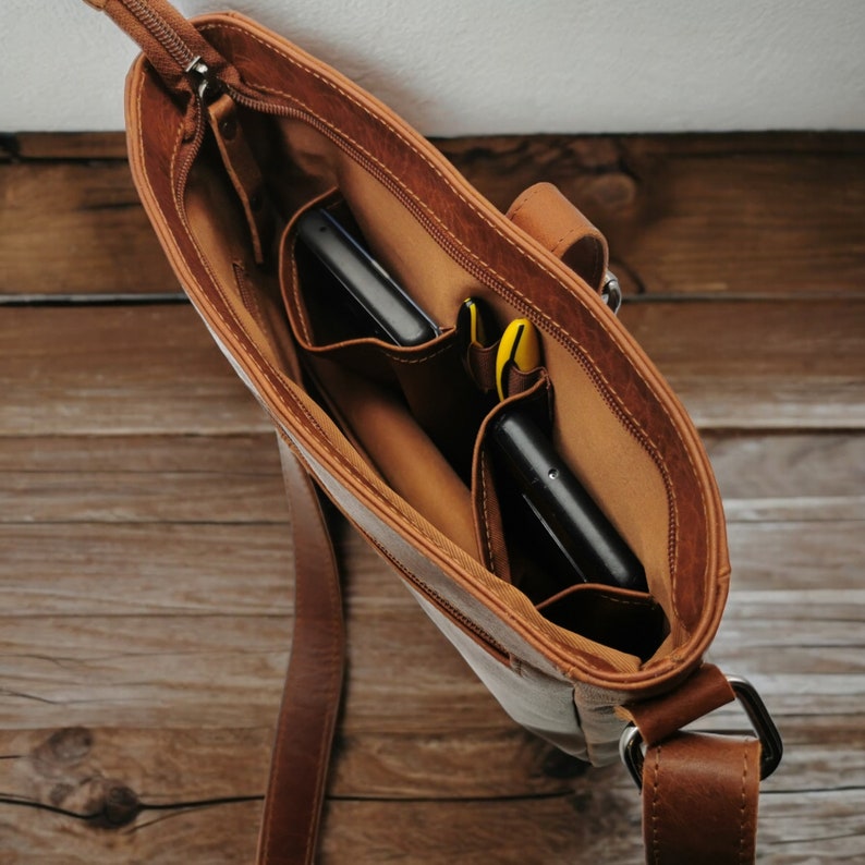 May include: A brown leather crossbody bag with a zipper closure and two interior pockets. The bag is open and shows two black phones and yellow pens inside.