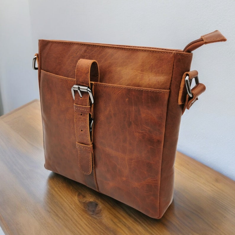 May include: A brown leather satchel bag with a silver buckle and a strap. The bag has a flap closure and a top handle.