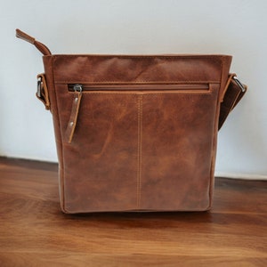 May include: A brown leather messenger bag with a zippered pocket and a flap closure. The bag has a long adjustable strap.