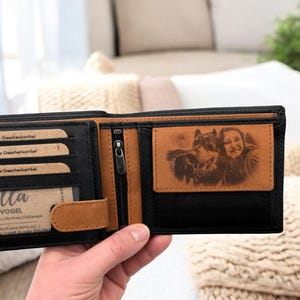 May include: A black leather wallet with a brown leather flap and a zippered pocket. The wallet has a personalized photo of a person and a dog engraved on the inside.