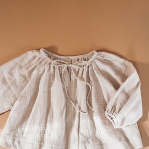 Pode incluir: Uma blusa de algod&atilde;o bege claro com decote franzido e um la&ccedil;o na frente. A blusa tem mangas compridas com punhos el&aacute;sticos.