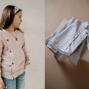 Könnte beinhalten: Zwei karierte Hemden, eines rosa mit braunen Knöpfen und eines weiß mit schwarzen Knöpfen. Beide Hemden haben lange Ärmel und eine lockere Passform.