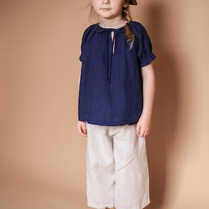 Pode incluir: Uma pessoa jovem vestindo uma blusa azul marinho com um la&ccedil;o no pesco&ccedil;o, cal&ccedil;as brancas e botas de tornozelo de couro marrom. A pessoa est&aacute; usando um bon&eacute; marrom.