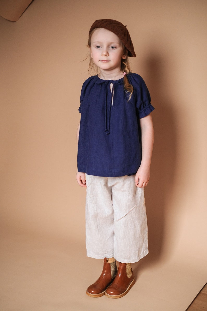 Pode incluir: Uma pessoa jovem vestindo uma blusa azul marinho com um la&ccedil;o no pesco&ccedil;o, cal&ccedil;as brancas e botas de tornozelo de couro marrom. A pessoa est&aacute; usando um bon&eacute; marrom.